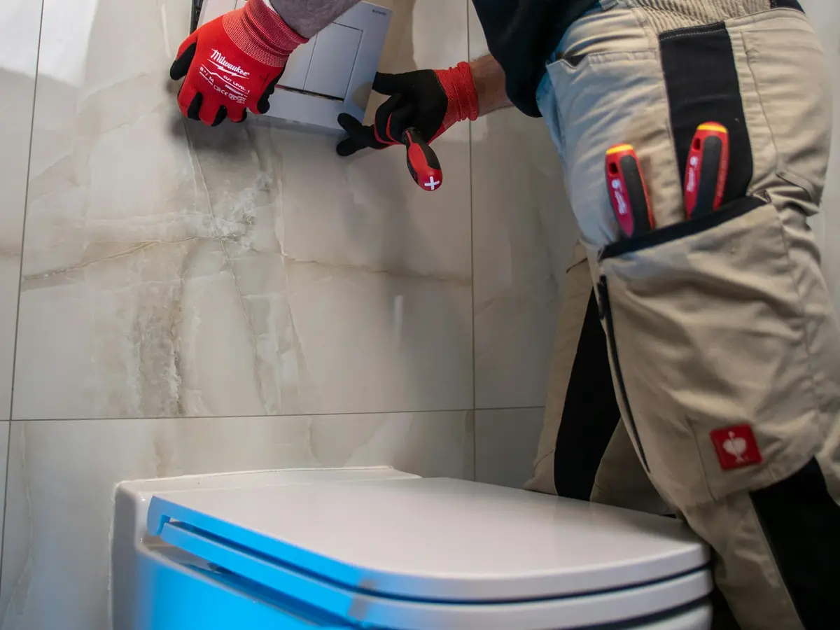 A plumber in red gloves working on the flush plate above a wall-mounted toilet